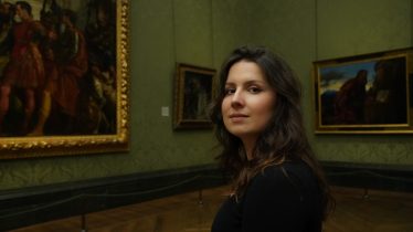 A woman with long wavy brown hair, standing in an art gallery, her face turned to look at the camera.